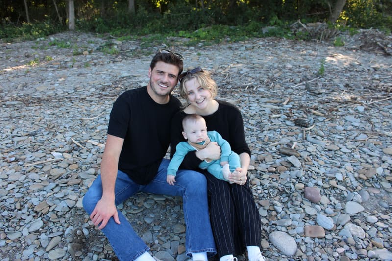 Jake and Danielle sitting together on a pebble beach, smiling, with Danielle holding their baby on her lap